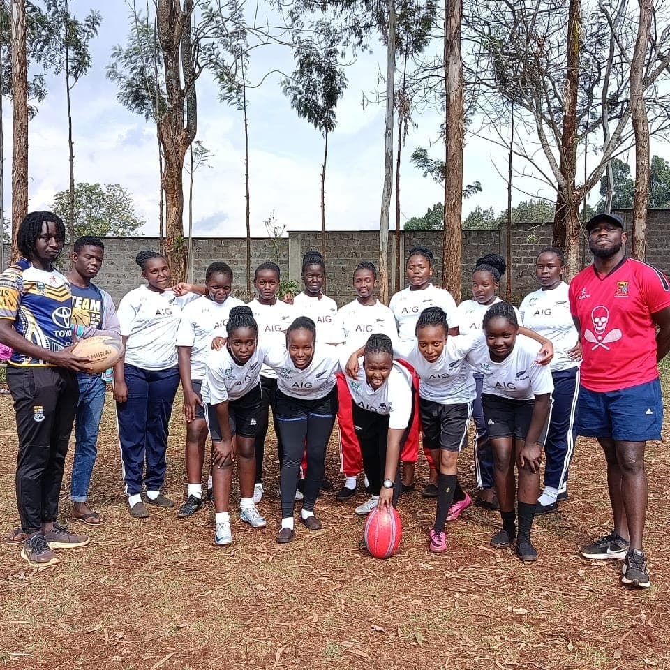 The girls' squad in white match jerseys gathered in two rows with their coaches, a rugby ball on the ground in front of them.